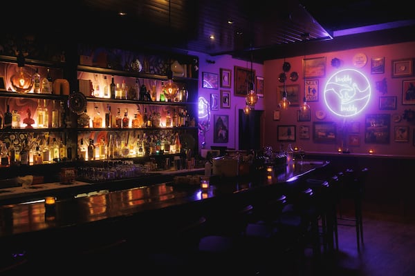 A darkly lit bar with lots of art on the walls and a pink neon sign of two hands reaching towards each other framed by the words buddy buddy