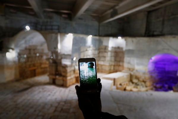 A worker holds up a cell phone displaying a photo of the Ein el-Fijeh reservoir, now seen dry in the background, in the Barada Valley, Syria, Thursday, May 8, 2025. The Ein el-Fijeh spring, the main water source for Damascus, has nearly dried up, forcing many residents in the capital and its suburbs to rely on water delivered by tanker trucks that fill from local wells. (AP Photo/Omar Sanadiki)
