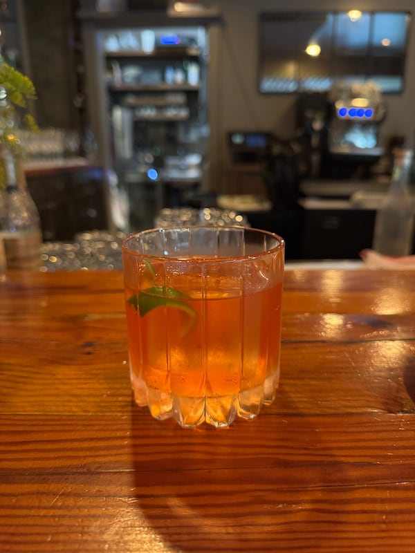A rose-hued cocktail in a rocks glass containing one large square of ice, garnished with a slice of lime peel.