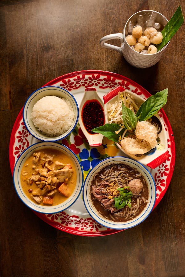 Overhead view of a tray of Thai food, including a bowl of curry, a bowl of noodle soup, a bowl of rice, a dish of sauce and a plate of herbs and sprouts.