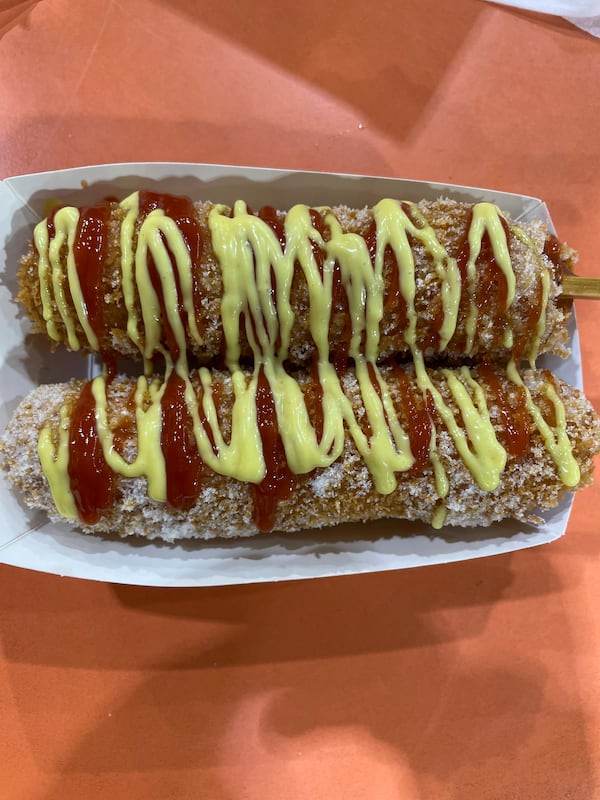 Two bunless hotdogs that have been battered in a rice-based batter and fried. They are drizzled with two sauces and placed in a paper takeout container.