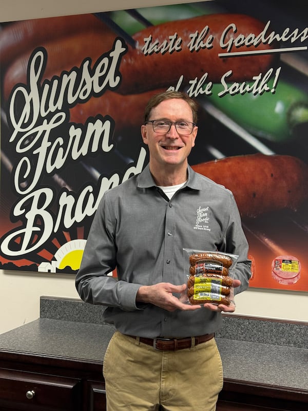 A man holds a package of sausage standing in front of a wall decorated to say Sunset Farm Foods