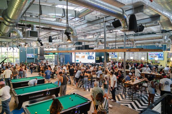 A busy, warehouse-sized bar filled with people, with pool tables on the left and large screen showing sports on the far wall