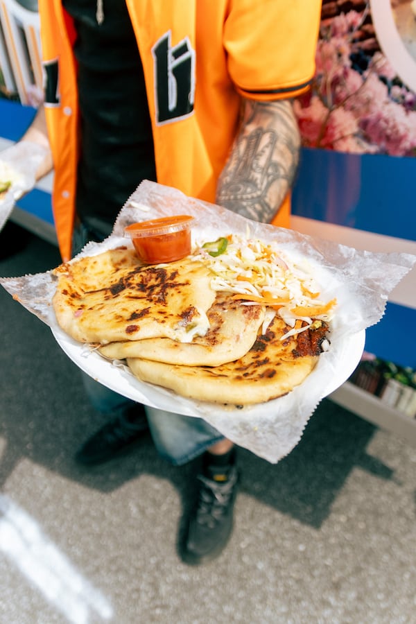 A man standing outside on a street holds a plate of three pupusas toward the camera.