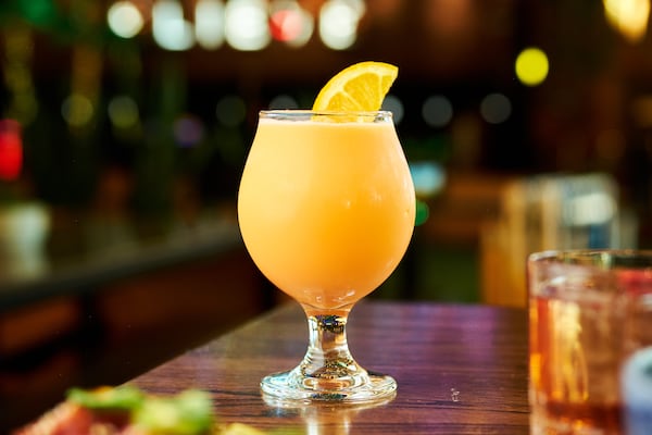 A brightly colored frozen cocktail is garnished with a half an orange slice. It's placed on the corner of a wooden table. There are out-of-focus lights and shapes that indicate the table is in front of a bar.