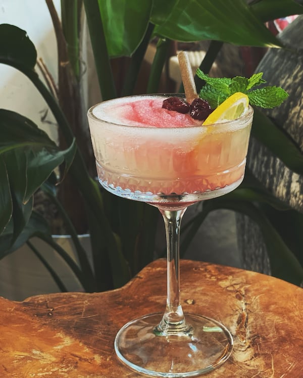 A cocktail glass with a long stem holds a frozen drink containing swirls of bright colors, bright fruit garnishes and a sprig of mint. It is placed on a table in front of large, lush plant leaves.