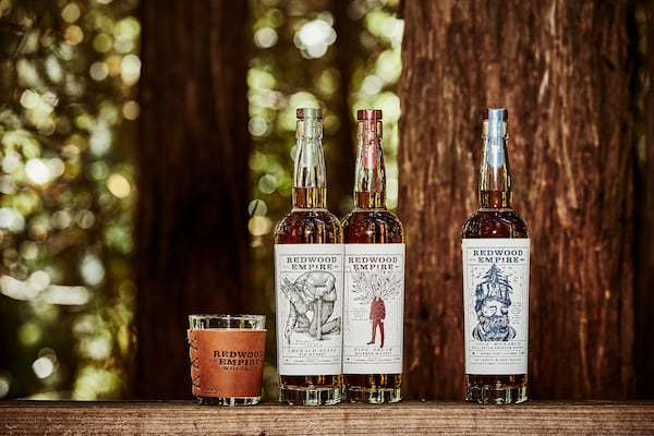 Bottles of Redwood Empire rye, bourbon and whiskey are lined up on a wood plank next to a tree, with  more trees in the background. A rocks glass featuring with the distillery name, Redwood Empire Distillery, is next to them.