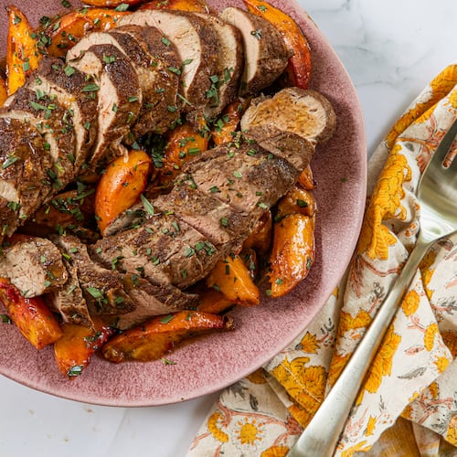 A plate shows two sliced pork tenderloins surrounded by roasted peach slices. A napkin with a print that complements the peaches' color is tucked next to the plate, and a serving fork rests on top of the napkin.