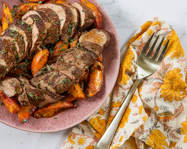 A plate shows two sliced pork tenderloins surrounded by roasted peach slices. A napkin with a print that complements the peaches' color is tucked next to the plate, and a serving fork rests on top of the napkin.
