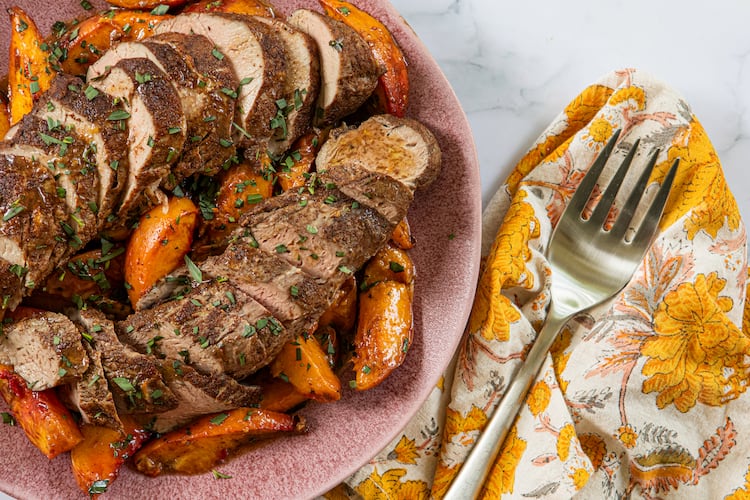 A plate shows two sliced pork tenderloins surrounded by roasted peach slices. A napkin with a print that complements the peaches' color is tucked next to the plate, and a serving fork rests on top of the napkin.