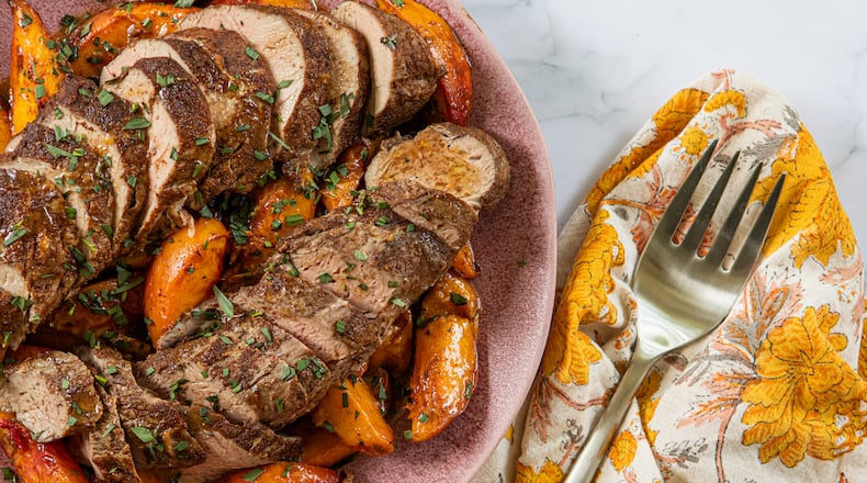 A plate shows two sliced pork tenderloins surrounded by roasted peach slices. A napkin with a print that complements the peaches' color is tucked next to the plate, and a serving fork rests on top of the napkin.