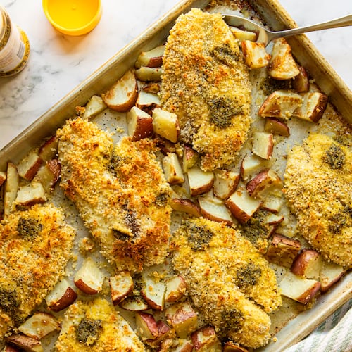 Six panko-coated tilapia fillets are on a sheet pan, with roasted red potato wedges scattered between them. A serving is lifting a fish fillet slightly. An open bottle of lemon pepper seasoning and a kitchen towel are also shown.