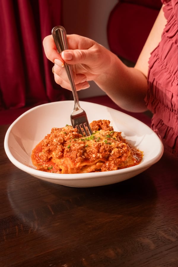 A woman's hand with a fork reaches over a plate of lasagna with extra tomato sauce.