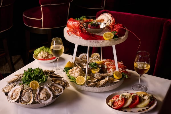 A seafood tower with lobster and oysters sits on a white tablecloth surrounded by more plates of chilled seafood, an heirloom tomato salad and a glass of white wine.