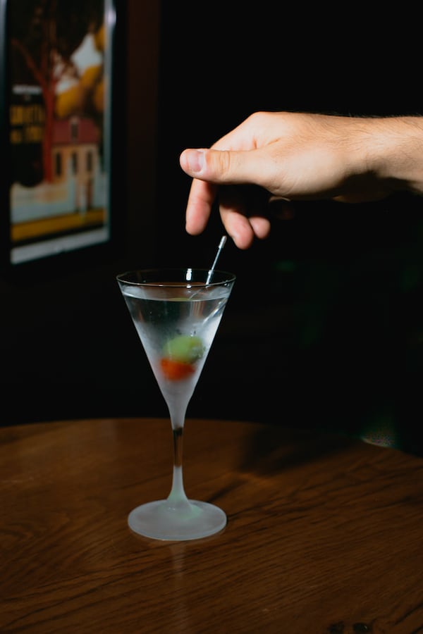 A martini sits on a wooden table while a hand stirs the drink.
