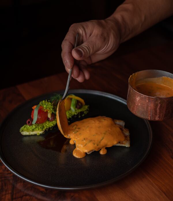 A hand with a spoon ladles a bright sauce over a grouper filet that contrasts with the dark plate