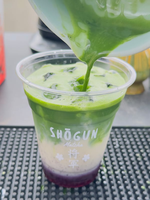 A blender is pouring matcha tea into a clear plastic cup. The cup has the Shogun Matcha logo on it, and is resting on a metal tabletop. The beverage has three layers:  a ube-colored layer on the bottom, a creamy looking middle layer, and the green matcha tea on top.