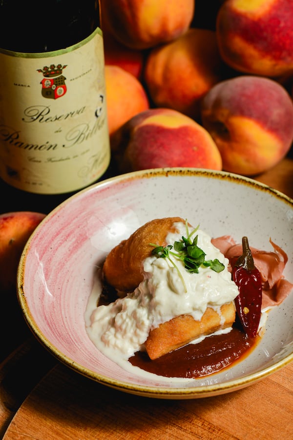 A dessert of peaches with whipped cream in a bowl next to a bottle of wine with a pile of peaches behind it.