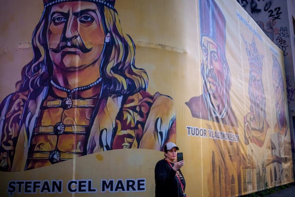 A woman holds up a mobile phone backdropped by depictions of Romanian historical figures outside the headquarters of the Alliance for the Unity of Romanians (AUR) shortly before polls close in the first round of the country's presidential election redo in Bucharest, Romania, Sunday, May 4, 2025. (AP Photo/Andreea Alexandru)