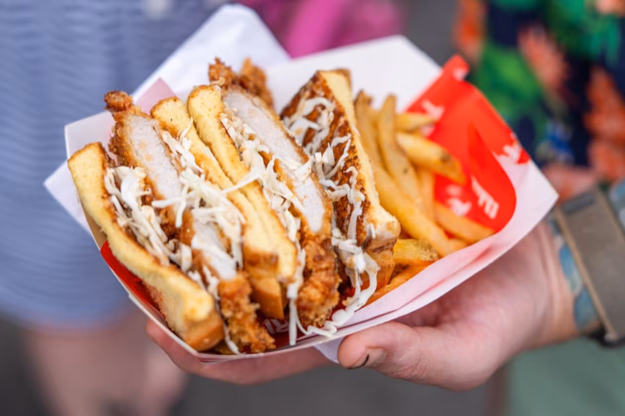 A hand holds Tori Atl’s katsu sando in a paper serving boat. The sandwich is sliced in half, showing the meat inside, and is served with french fries.