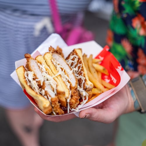 A hand holds Tori Atl’s katsu sando in a paper serving boat. The sandwich is sliced in half, showing the meat inside, and is served with french fries.