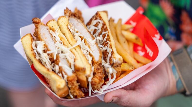 A hand holds Tori Atl’s katsu sando in a paper serving boat. The sandwich is sliced in half, showing the meat inside, and is served with french fries.