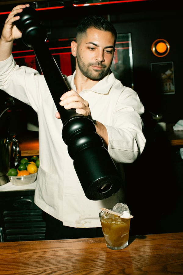 A man holds an oversized black pepper grinder over a cocktail as he cracks pepper on it.