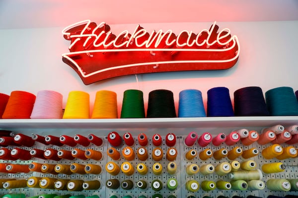 A fluorescent Huckmade logo hangs above spools of colorful sewing threads at Huckleberry Starnes’ studio in Decatur on Monday, April 27, 2026. (Miguel Martinez/AJC)