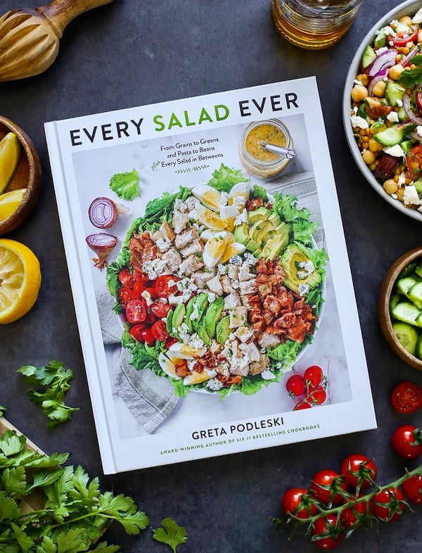 a cookbook titled Every Salad Ever, featuring a bowl of salad with lettuce, avocado, tomato, egg and proteins on a dark table surrounded by herbs and produce