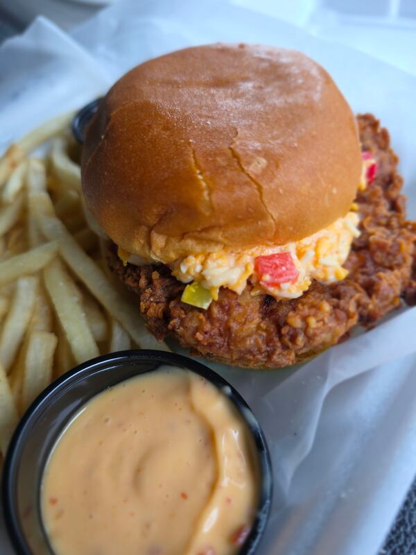 A fried porch chop sandwich on a brioche bun topped with a signature sauce. There are french fries on the side, plus a little cup of additional sauce.