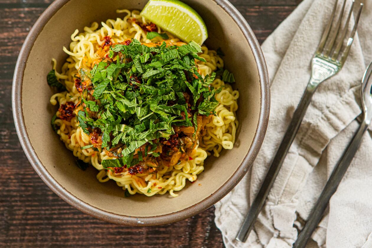 A bowl of ramen noodles rests on a wooden table. The color of the bowl complements the color of the wood. On top of the noodles is a chili crisp zucchini sauce, which is also a color that resembles the wood and bowl. On top of the sauce is a mound of fresh mint leaves, whose color is a bright, happy contrast. Also on the table is a napkin, fork and knife.
