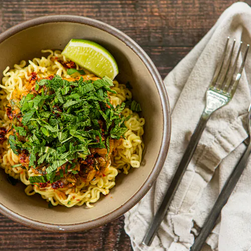 A bowl of ramen noodles rests on a wooden table. The color of the bowl complements the color of the wood. On top of the noodles is a chili crisp zucchini sauce, which is also a color that resembles the wood and bowl. On top of the sauce is a mound of fresh mint leaves, whose color is a bright, happy contrast. Also on the table is a napkin, fork and knife.
