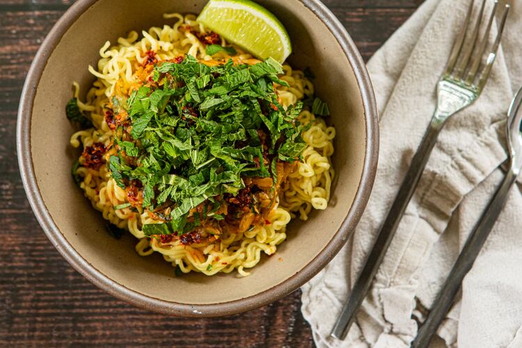 A bowl of ramen noodles rests on a wooden table. The color of the bowl complements the color of the wood. On top of the noodles is a chili crisp zucchini sauce, which is also a color that resembles the wood and bowl. On top of the sauce is a mound of fresh mint leaves, whose color is a bright, happy contrast. Also on the table is a napkin, fork and knife.