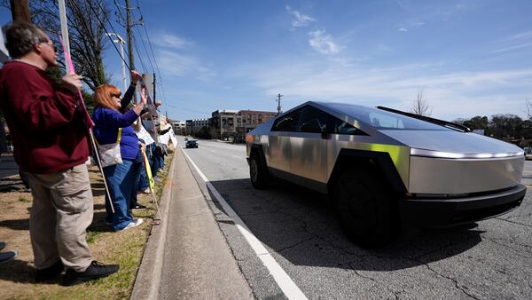 People stand with other demonstrators as a Cybertruck drives by during a protest of Elon Musk near a Tesla vehicle dealership on Saturday, March 8, 2025, in Decatur. (Mike Stewart/AP)