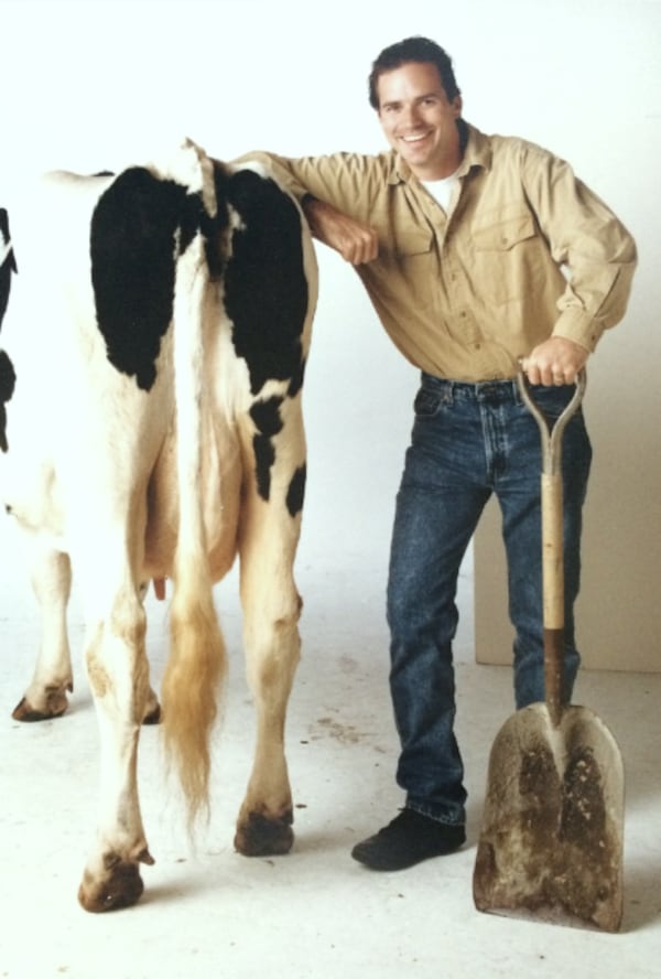 A cow stands with its behind facing the camera, and David Ring stands smiling beside the cow with an arm on its back and a shovel in his other hand.