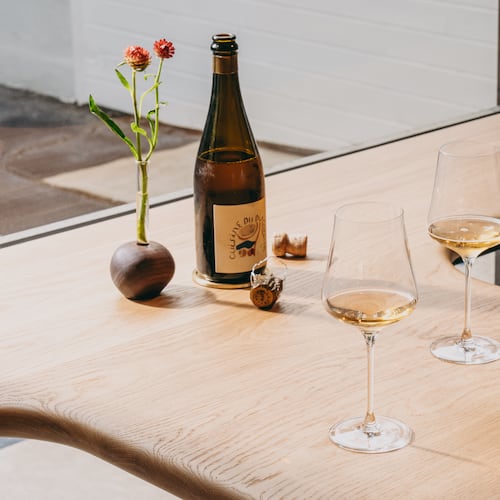 A smooth, light wooden counter sits against a bright window. On the counter, there is a little bouquet of flowers, a wine bottle and two half-filled wine glasses.