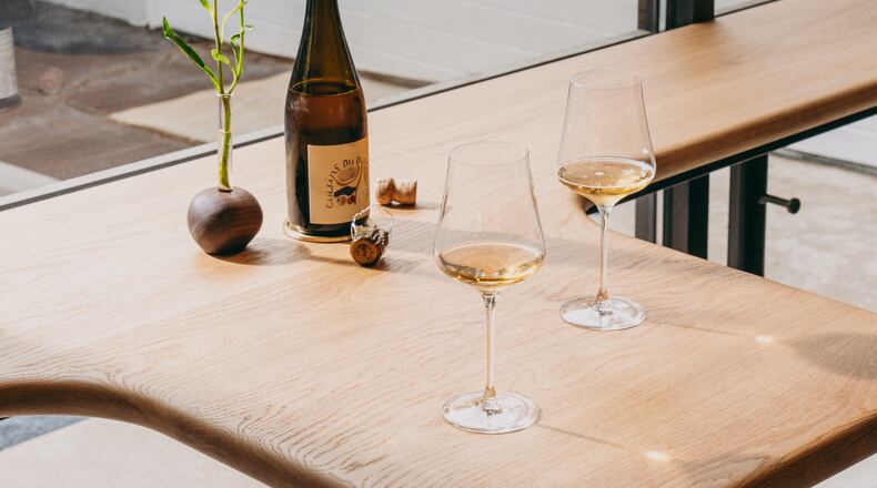 A smooth, light wooden counter sits against a bright window. On the counter, there is a little bouquet of flowers, a wine bottle and two half-filled wine glasses.