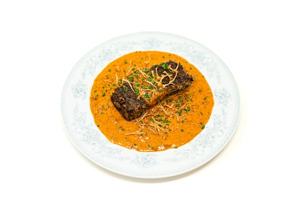A very stylized image of a wedge of meatloaf in a pool of sauce, garnished with specks of green and black herbs and enoki mushrooms.