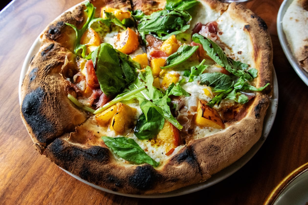 A whole pizza, with a thick, charred crust, is topped with cheese, peaches, prosciutto and ribbons of fresh arugula.