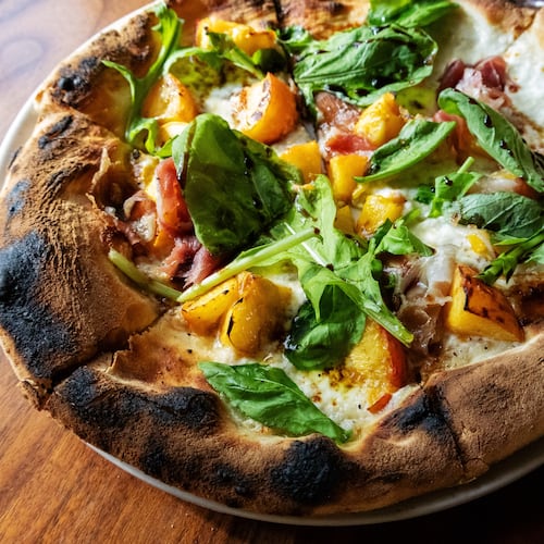 A whole pizza, with a thick, charred crust, is topped with cheese, peaches, prosciutto and ribbons of fresh arugula.