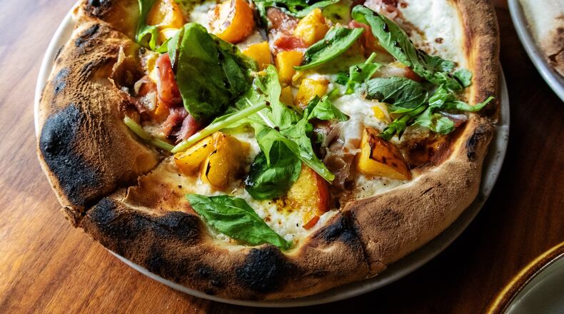 A whole pizza, with a thick, charred crust, is topped with cheese, peaches, prosciutto and ribbons of fresh arugula.