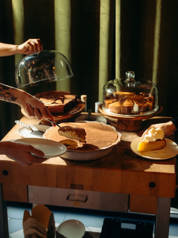 A wooden dessert cart where someone is serving tiramisu from a large bowl while another person lifts a glass cloche off of a chocolate cake. A lemon-olive oil cake sits in the back right.