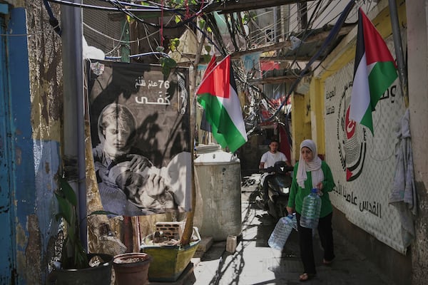 A Palestinian woman carries water containers through an alley decorated with Palestinian flags and posters marking the 76th anniversary of the Nakba, in the Mar Elias refugee camp in Beirut, Lebanon, Monday, June 2, 2025. The Arabic writing on the poster reads, "76 years of injustice, enough." (AP Photo/Hassan Ammar)