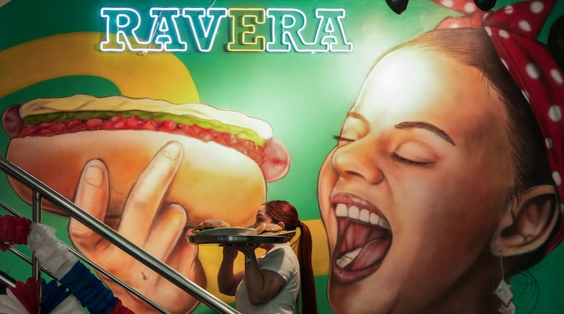 A waitress carries a tray with "completos", traditional Chilean hot dogs topped with mashed avocado, diced tomatoes and mayonnaise, past a mural dedicated to the dish at the Ravera restaurant in downtown Santiago, Chile, Thursday, Sept. 10, 2026. (AP Photo/Esteban Felix)