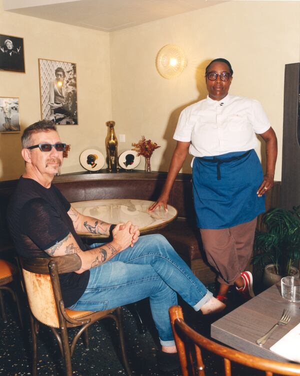 In a brightly lit restaurant dining room, a man sits in a bistro chair on th left side of the frame wearing sunglasses, while a woman who is a chef stands on the right while leaning slightly on a table.