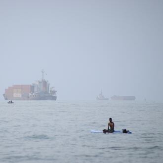 FILE - A man sits on a surfboard as a container ship and other commercial vessels appear anchored in the Strait of Hormuz off Bandar Abbas, Iran, Monday, July 27, 2026. (Razieh Poudat/ISNA via AP, File)