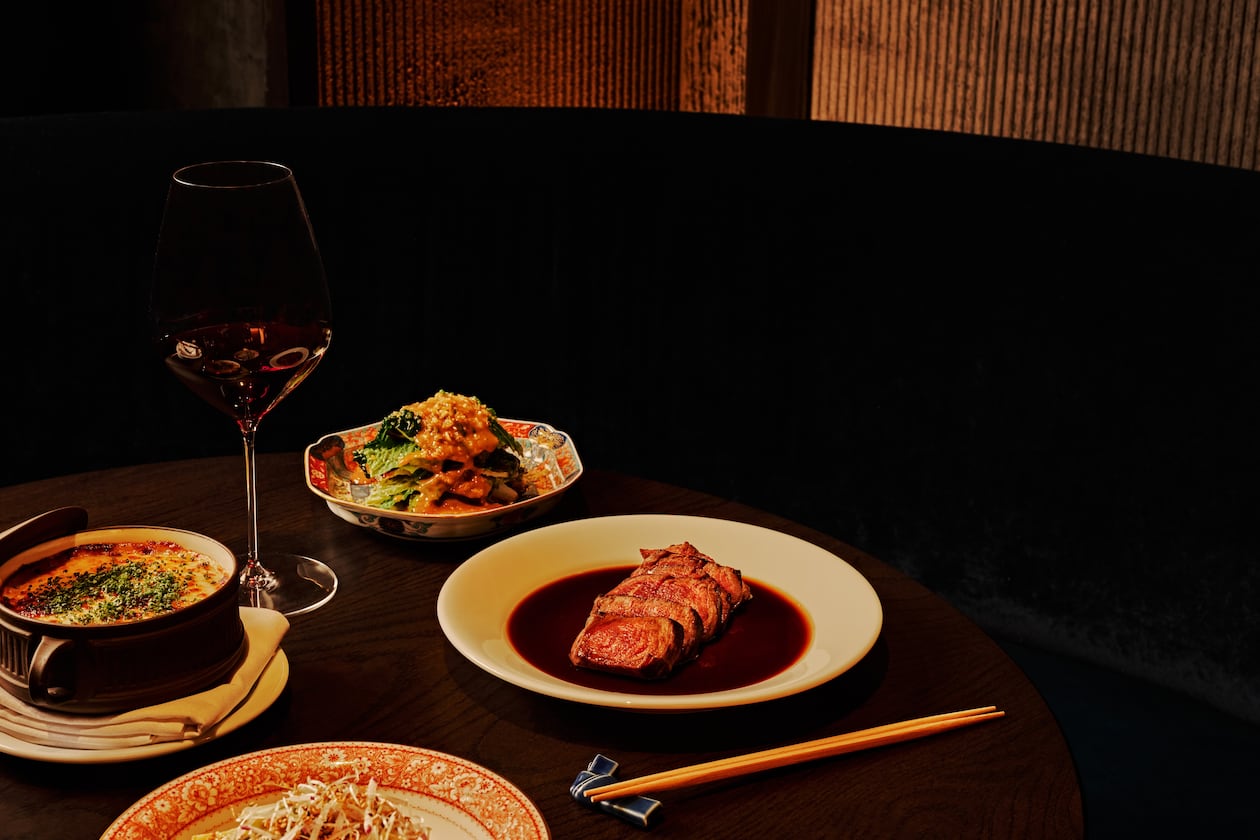 A selection of dishes, including a sliced filet mignon and three vegetable sides, sit on a table in a dark, moody room with a glass of wine