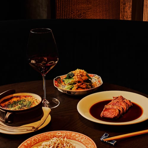 A selection of dishes, including a sliced filet mignon and three vegetable sides, sit on a table in a dark, moody room with a glass of wine