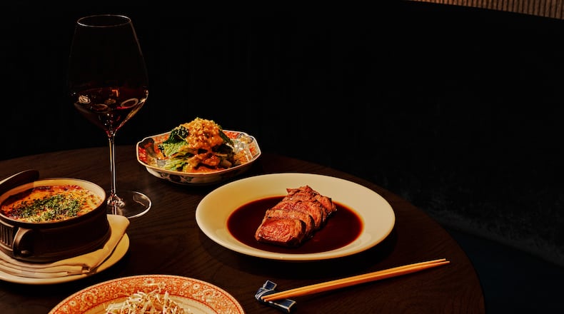 A selection of dishes, including a sliced filet mignon and three vegetable sides, sit on a table in a dark, moody room with a glass of wine