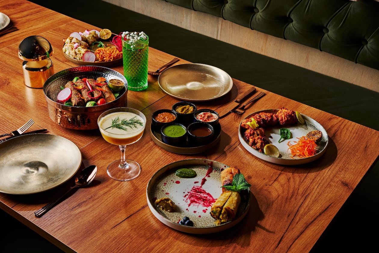 A group of several plates of indian food with colorful cocktails and gold place settings on a wooden table